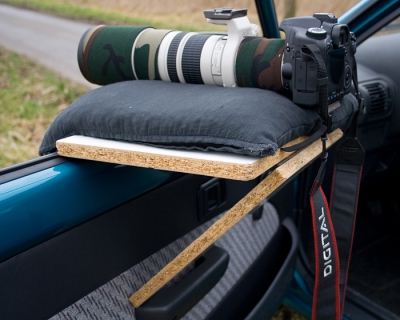 Deze staat ook in mijn pa op birdpix, maar kan ook voor nederpixers handig zijn. Oorspronkelijke ontwerper: Rein Hofman - birdfocus.nl
Benodigheden: 2 plankjes, 1 pianoschanier en 2 hoekijzertjes. De plankjes vooraf zoals getoond positioneren en zo op de juiste maat aftekenen. De breedte en lengte geheel naar eigen smaak en maat van rijstzak en telelens afstemmen. Hoekijzertjes kun je het beste aan de rechterkant (nabij buitenspiegel) plaatsen zodat ze niet het glas raken van je zijruit. De hoekijzertjes plaats je dus in de gleuf van je autoraam en zetten de rand van de dragende plank vast. Het onderste plankje zorgt ervoor dat de dragende plank aan de onderkant wordt 'schrap gezet'. De dragende plank wordt van 2 kanten vergrendelt, en kan dus geen milimeter verschuiven. Je kunt ook gerust met je lens erop een stukje verder rijden, zolang je geen diepe kuilen in rijdt blijft je lens mooi liggen. 

Nog een opmerking: het kan zijn dat de plank met 1 rijstzak erop te laag ligt om comfortabel te kunnen werken, je zou met je nek in de verrekking moeten gaan. Dit kun je het eenvoudigst oplossen door 2 rijstzakken op elkaar te leggen. Andere optie is om de onder de buitenrand van de dragende plank een balkje te timmeren en de schuine plank iets langer te maken. Zo komt de dragende plank hoger te staan. Wel natuurlijk vooraf goed passen en meten. 

Op foto de Canon EOS 40D & EF 400 mm f5.6 L USM